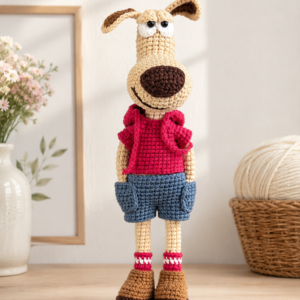 Crocheted long-nosed dog doll in a red hoodie and blue shorts, standing on a shelf beside a vase of flowers
