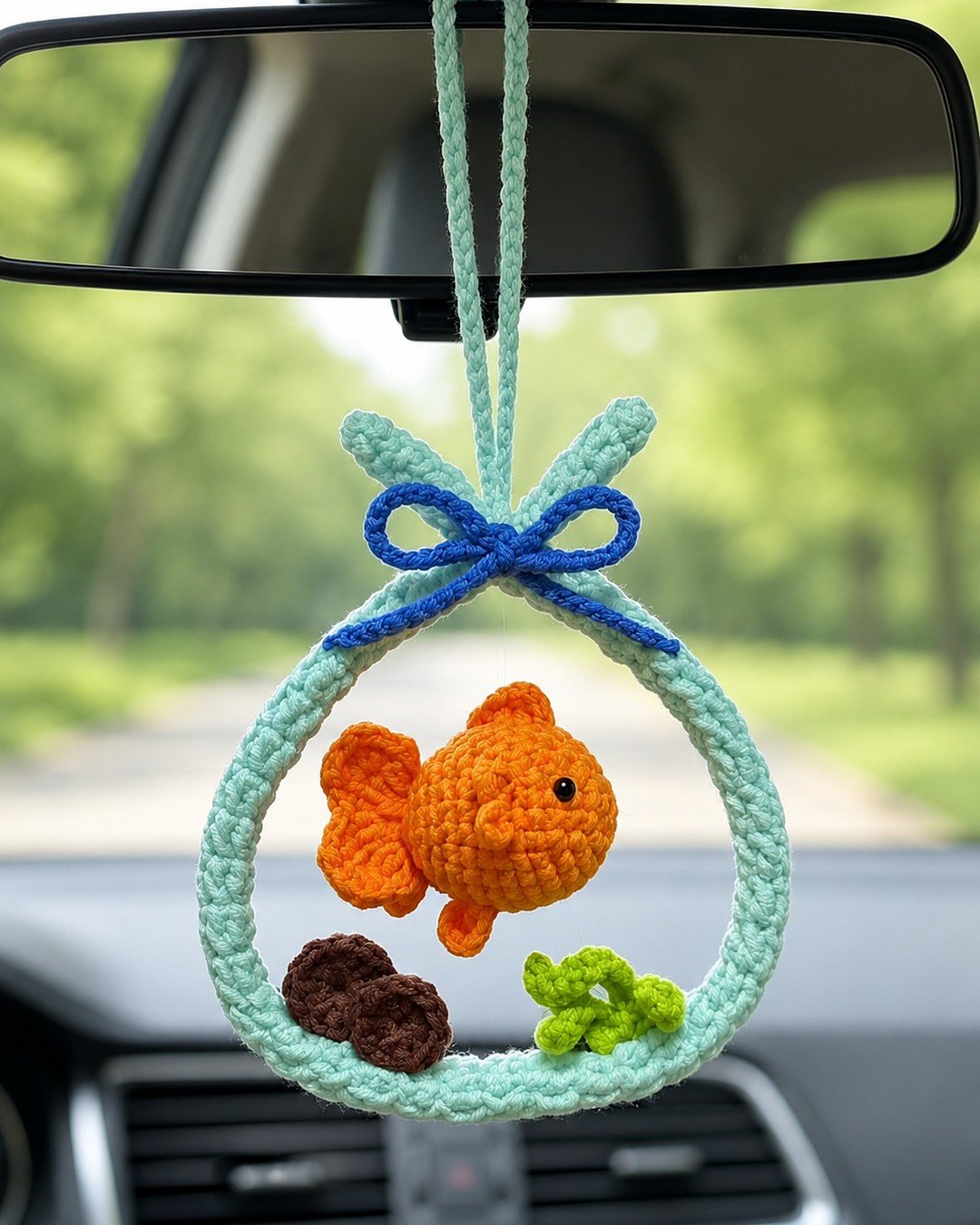 The goldfish aquarium ornament hanging from a car rear-view mirror with a tree-lined road ahead