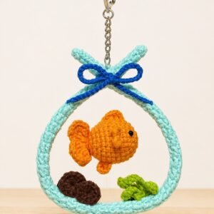 Crocheted goldfish ornament: an orange fish suspended inside a mint aquarium hoop with a blue bow, brown rocks and green leaves, on a keyring clasp