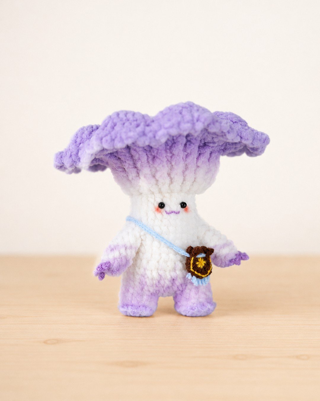 Crocheted white mushroom doll with a ruffled purple cap, standing on a wooden surface with a small brown crossbody bag