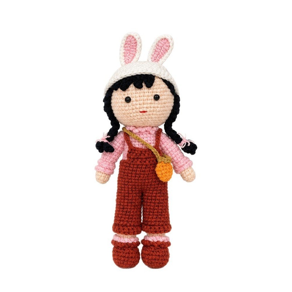 Wren, a crocheted doll in a white bunny-ear hat, pink sweater and brick-red overalls with an orange bag